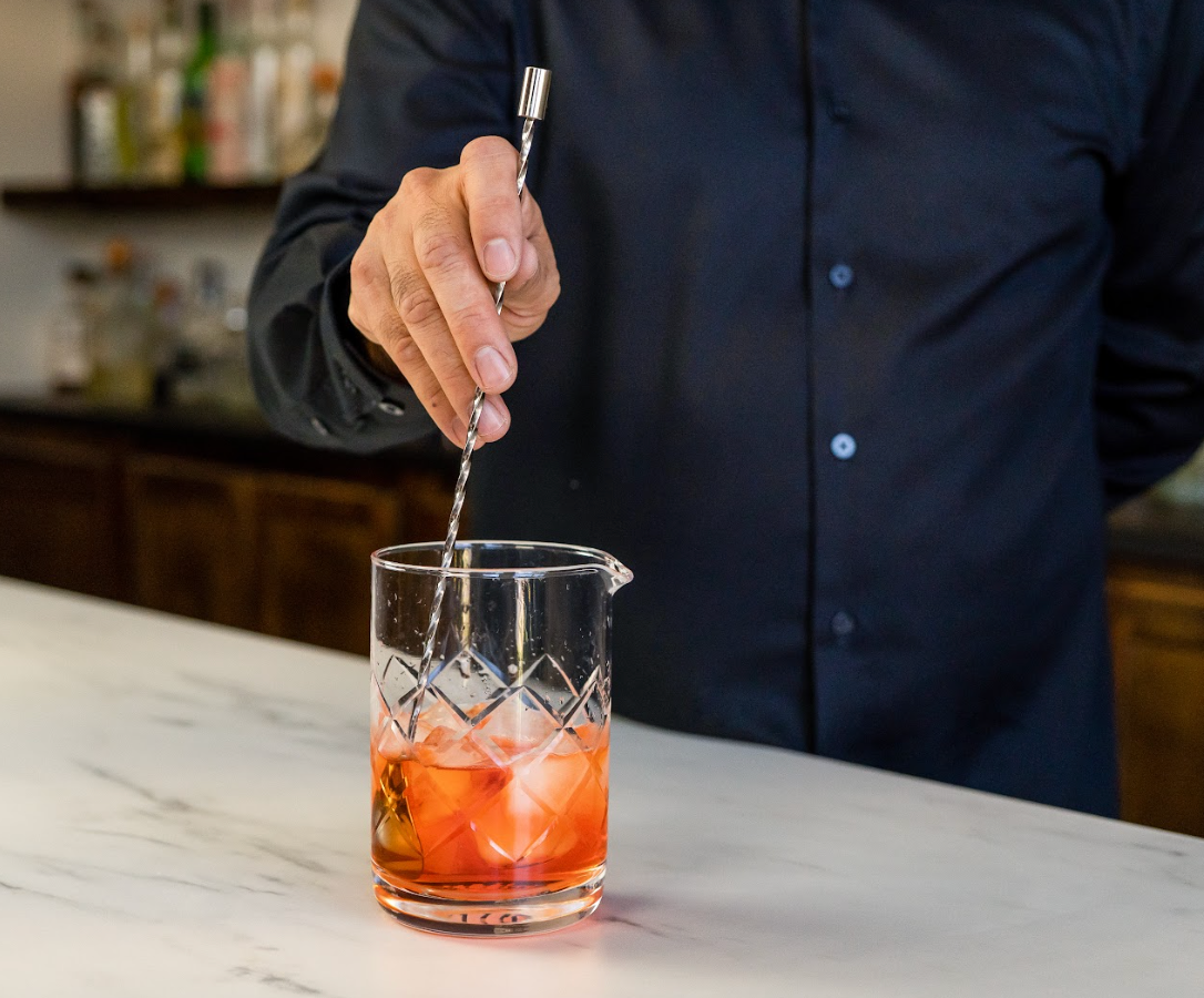 Hand stirring a red-orange cocktail in a mixing glass with a stainless steel bar spoon with spiral handle