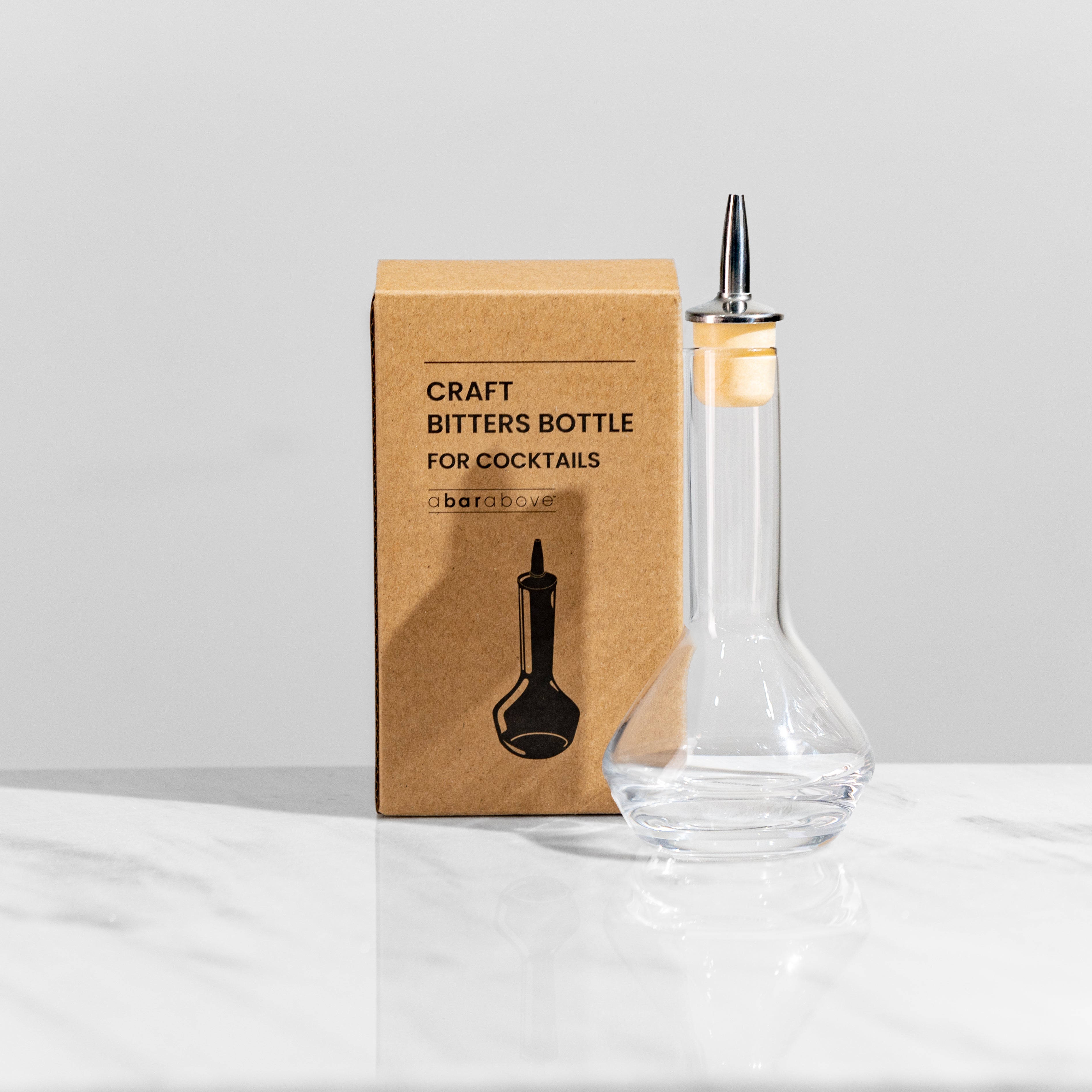 1 clear bitters bottle with rounded bottom on a white marble table, next to its gift box that reads "Craft bitters bottle for cocktails"