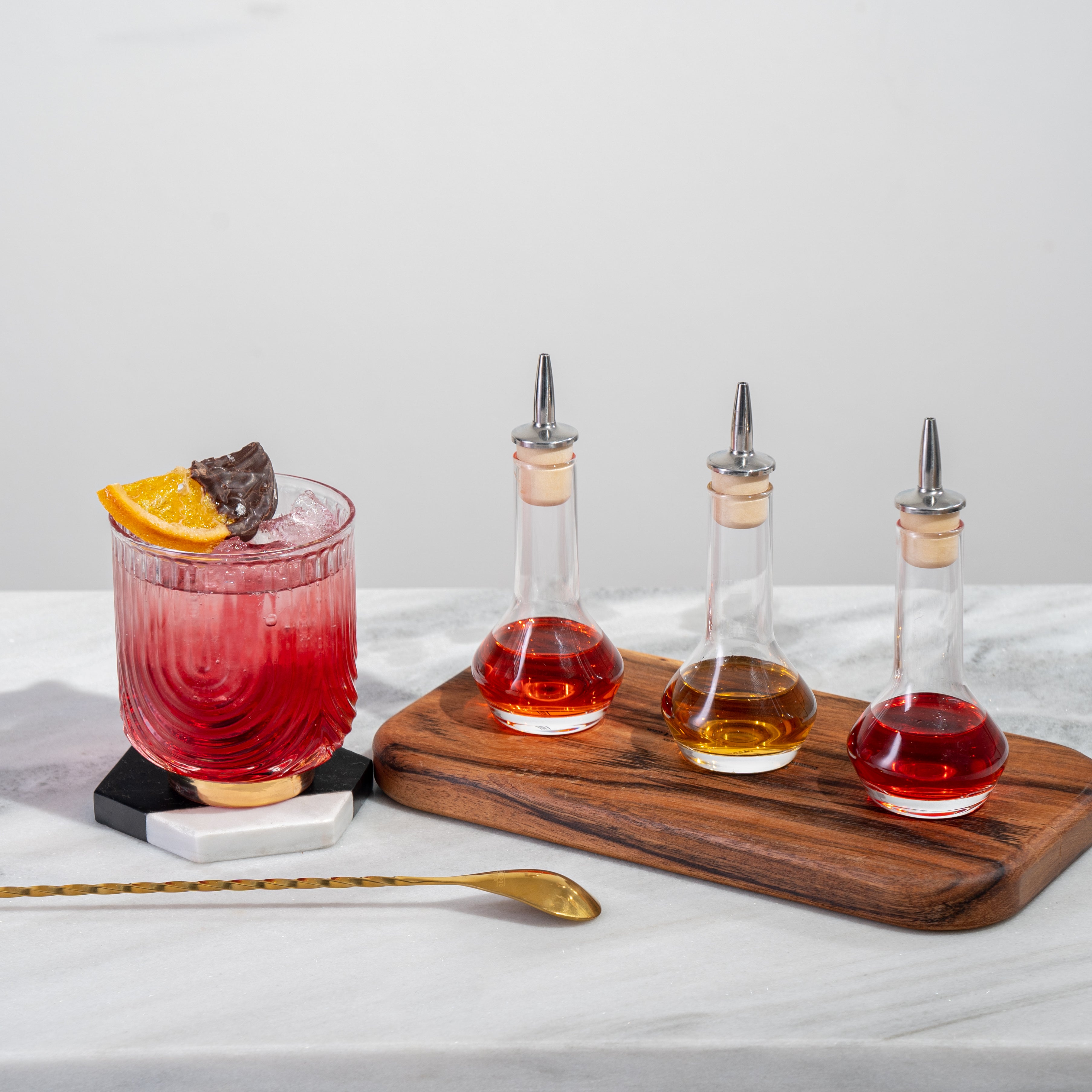 Red cocktail with candied orange garnish sitting next to 3 clear bitters bottles with red and amber liquid, next to a gold bar spoon