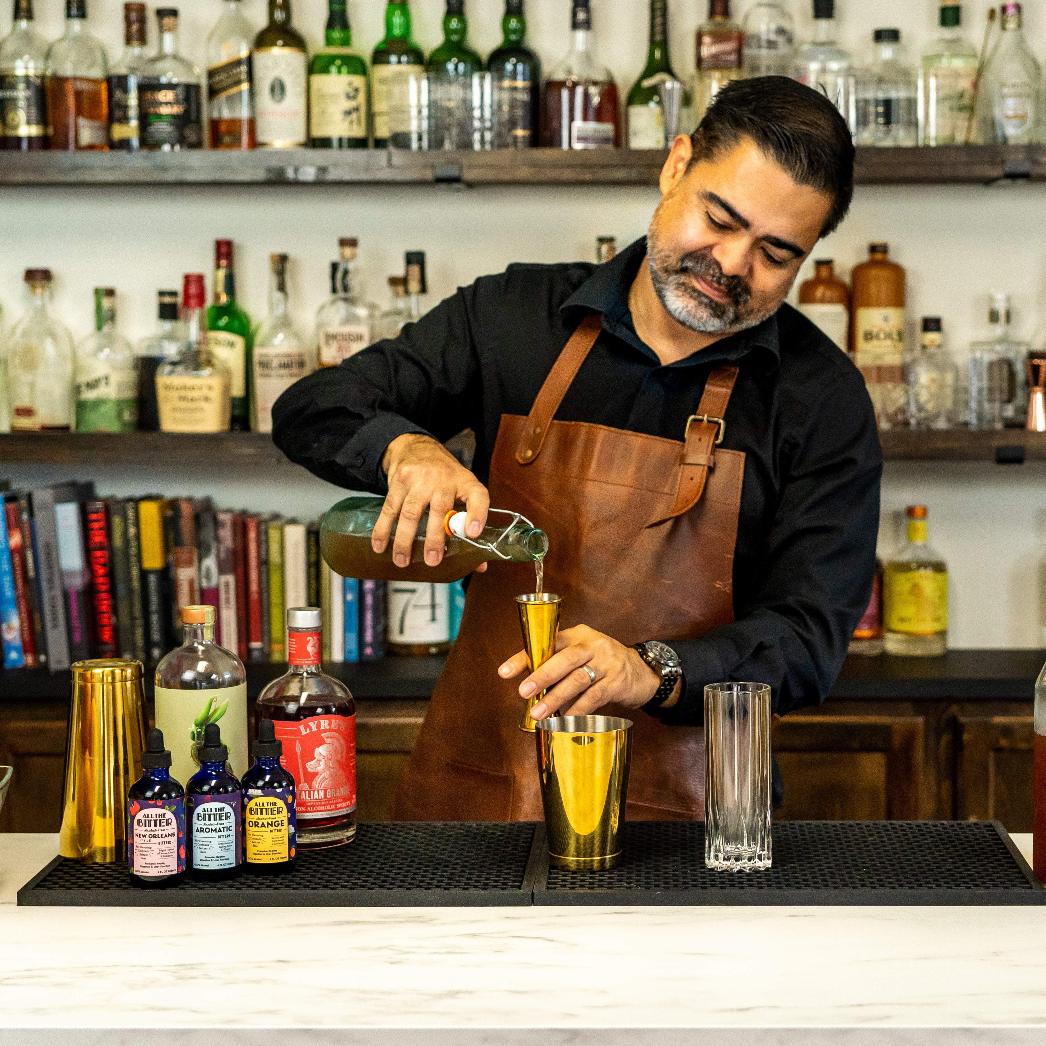 Bartender pouring syrup into a gold cocktail jigger at a bar, with bitters & mocktail ingredients, tall glass, & cocktail shaker on the bar