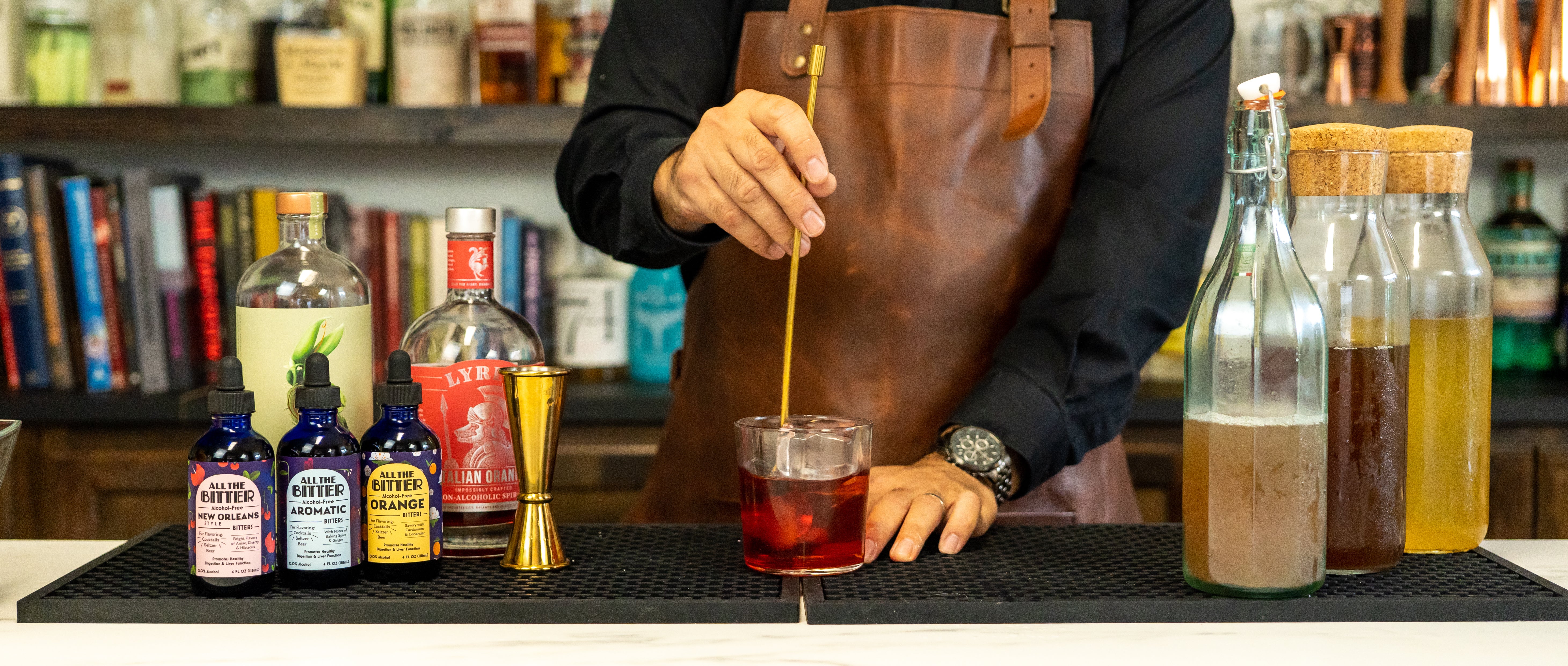 Bartender stirring cocktail in a rocks glass with a gold bar spoon, surrounded by cocktail bitters, bar tools, & non-alcoholic spirits