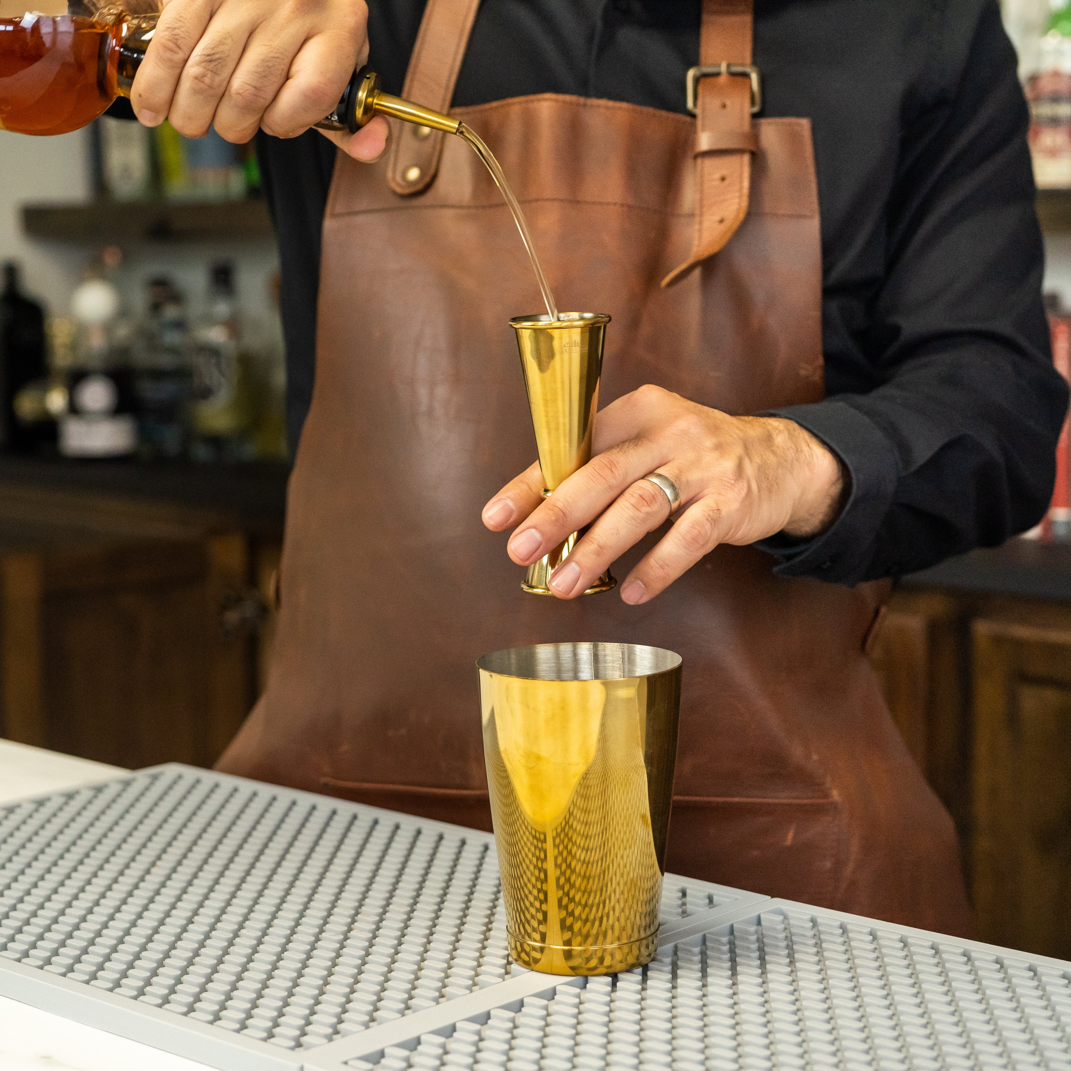 Bartender's hands pouring alcohol from a bottle with a pour spout into a gold cocktail jigger above a shaker tin