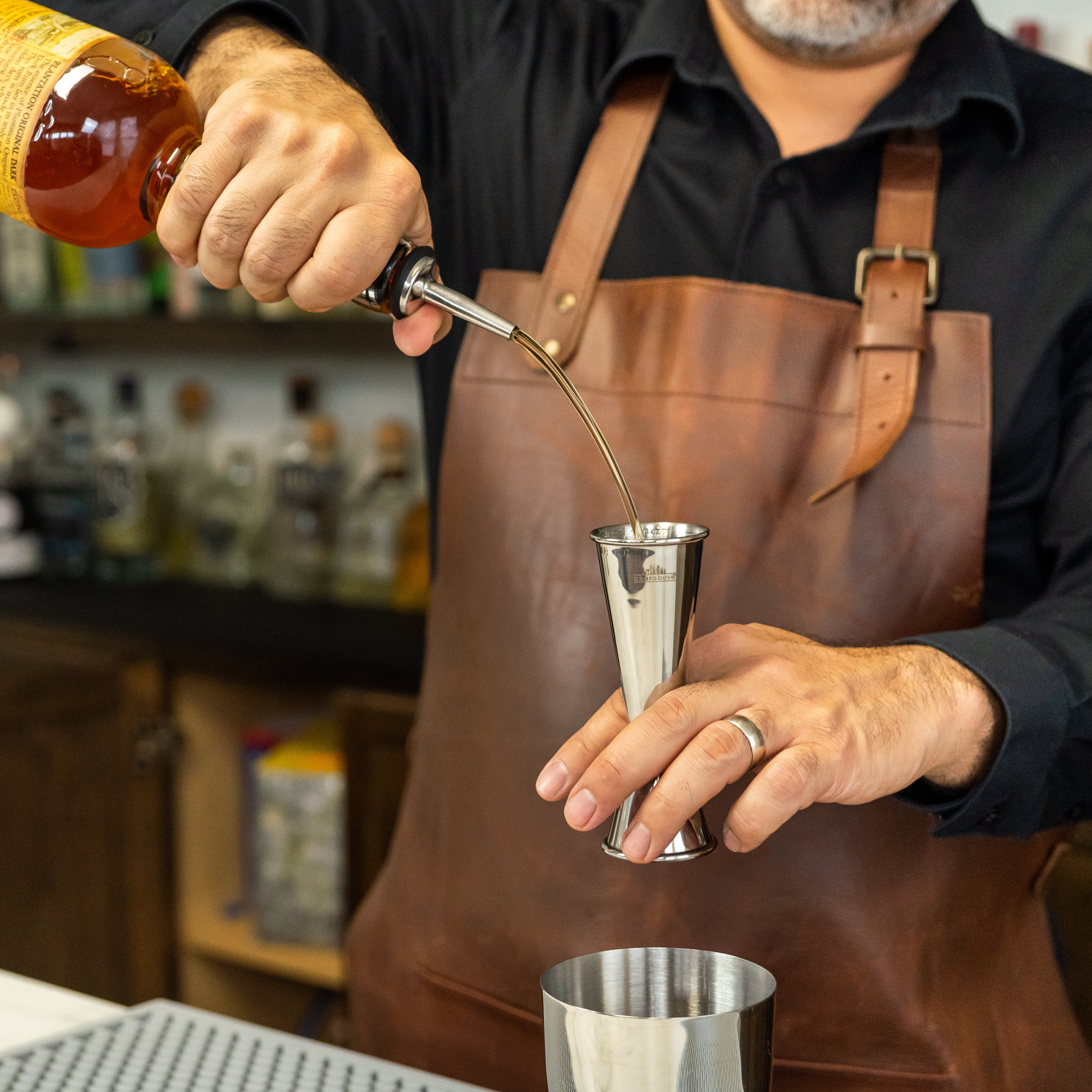 Bartender's hands pouring alcohol from a bottle with a pour spout into a stainless steel cocktail jigger above a shaker tin