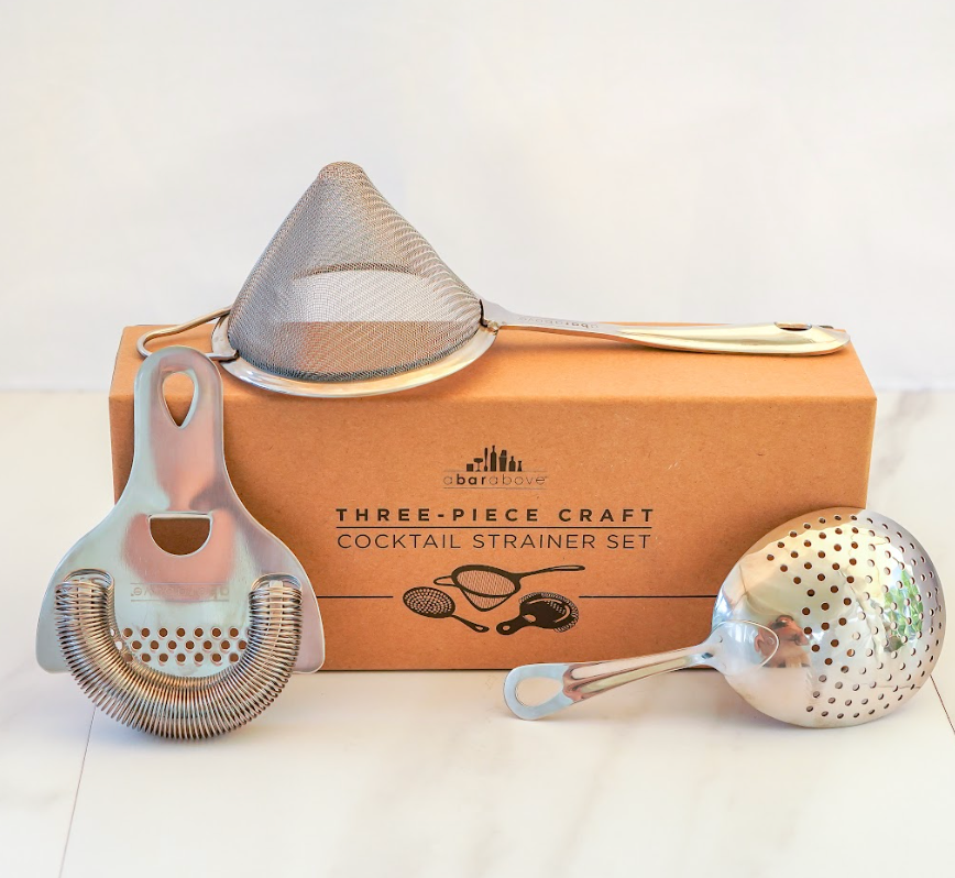 Gift box that says "three-piece craft cocktail strainer set" surrounded by Hawthorne, julep, and fine strainers on white marble