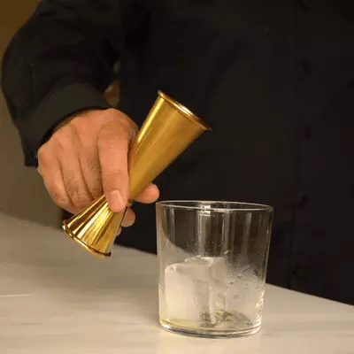 Hand pouring liquid from an bronze pour spout into a cocktail jigger behind a clear mixing glass with brown alcohol, in a pro bar