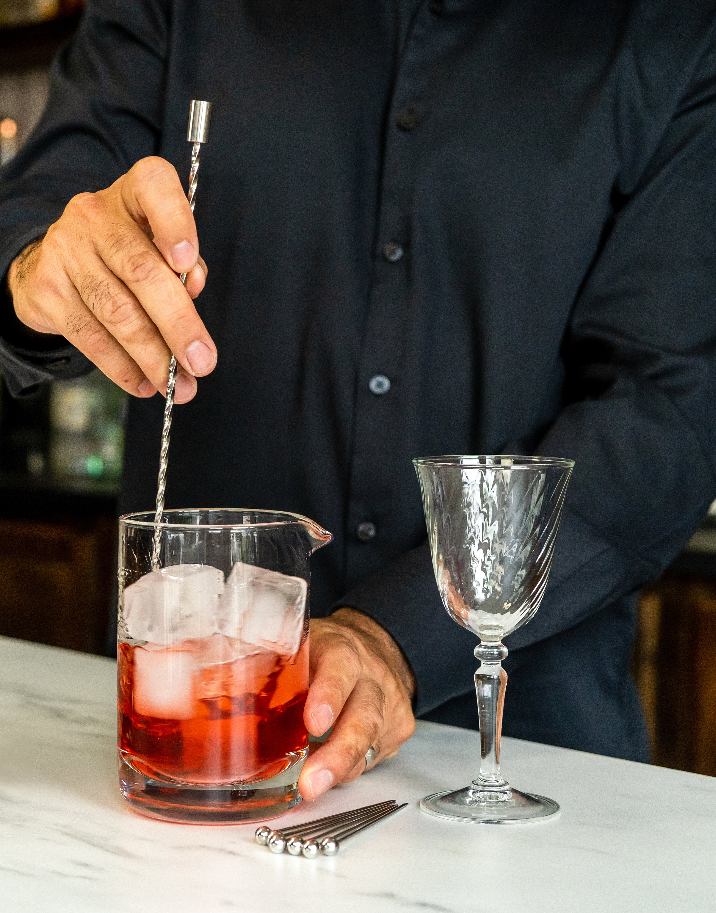 Hand mixing a red cocktail in mixing glass with ice, using a steel bar spoon. Cocktail picks & empty art deco wine goblet sit on the side