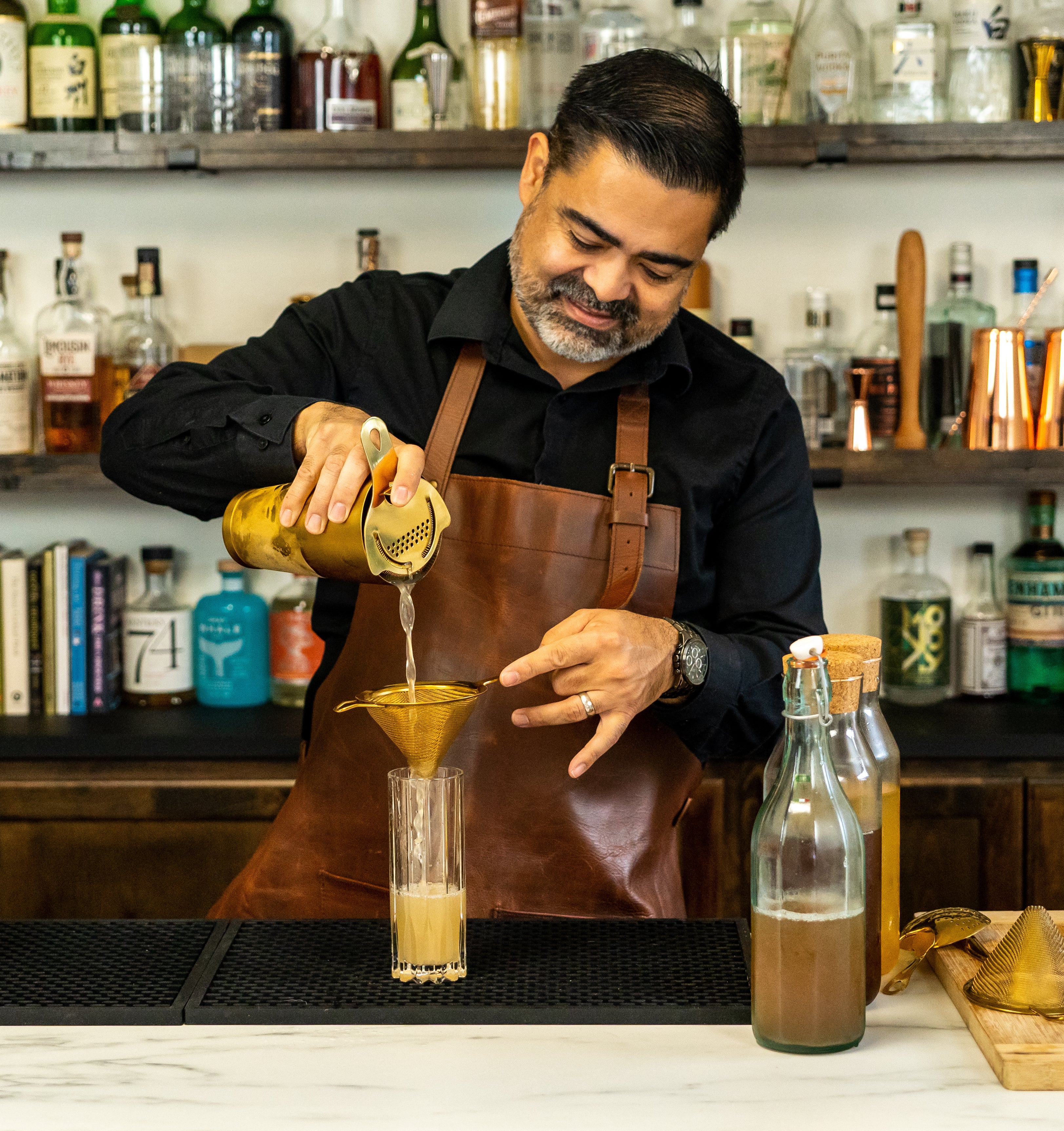 Bartender pouring brown cocktail from a gold cocktail shaker with Hawthorne strainer & fine strainer into a tall highball glass