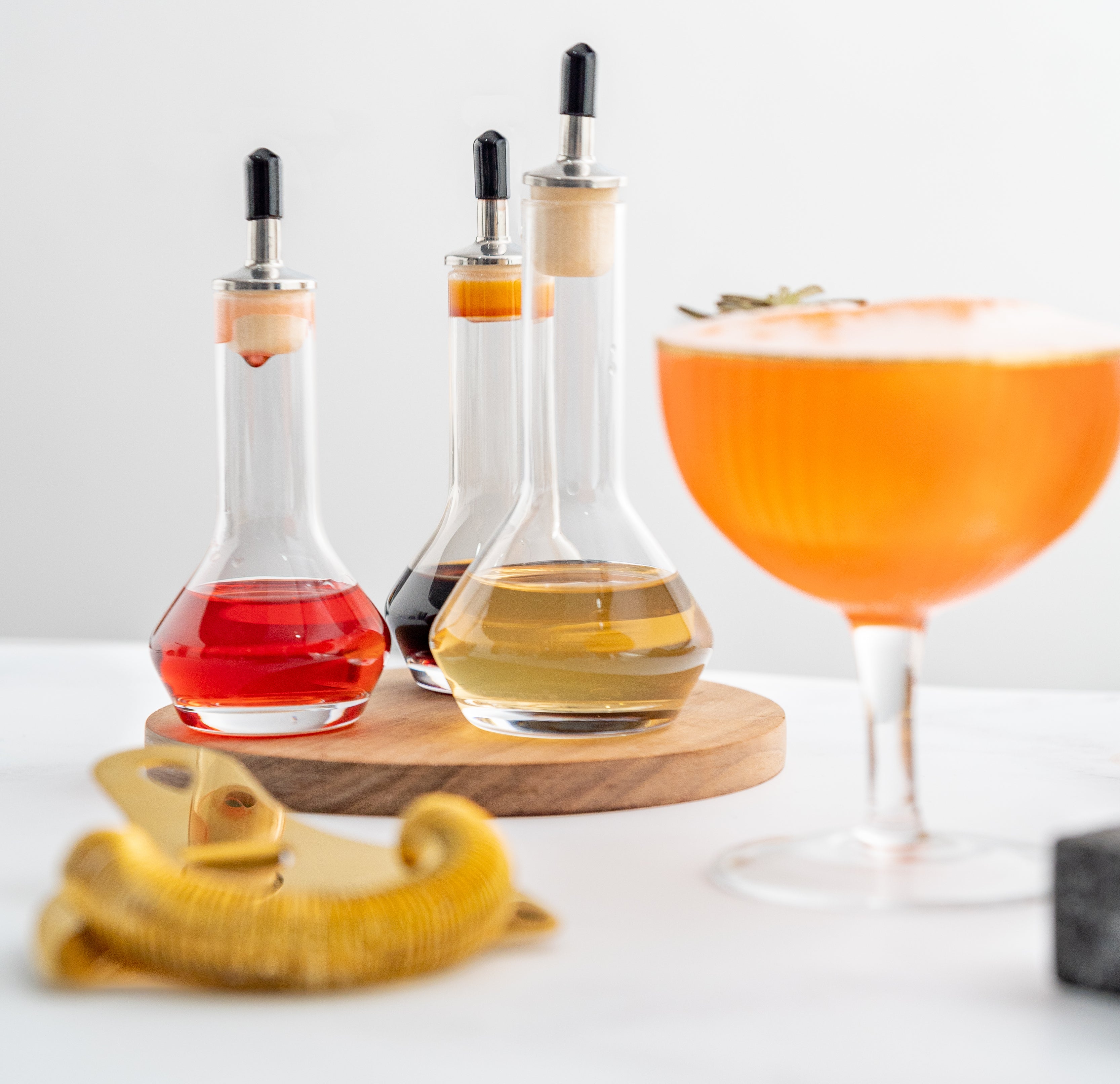 3 clear bitters bottles with round bottoms filled with liquid, with blurred out orange cocktail and gold strainer in the foreground