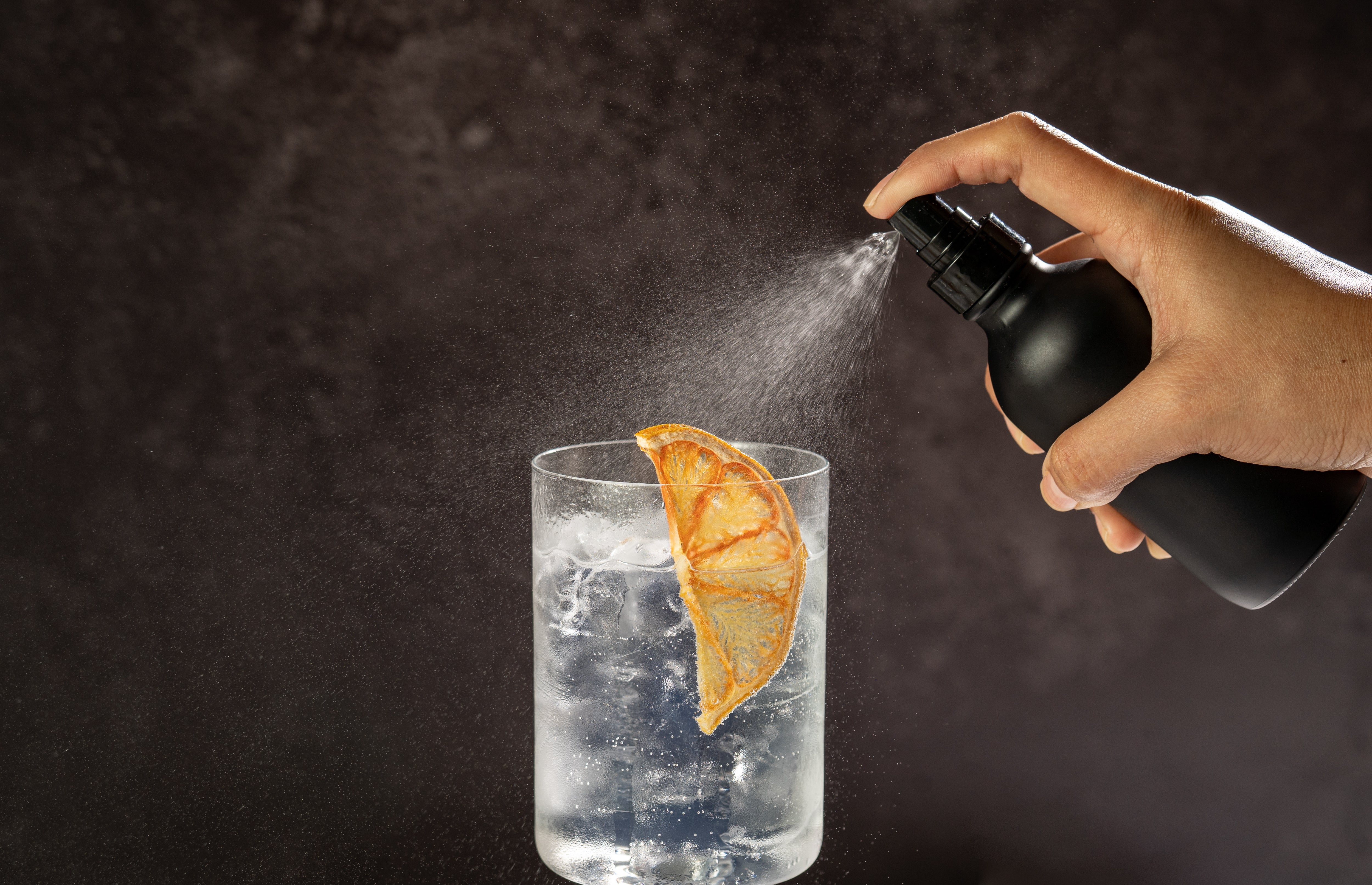 Hand spraying liquid from a black atomizer bottle into a rocks glass with vodka, ice, and dried orange garnish, against a brown backdrop