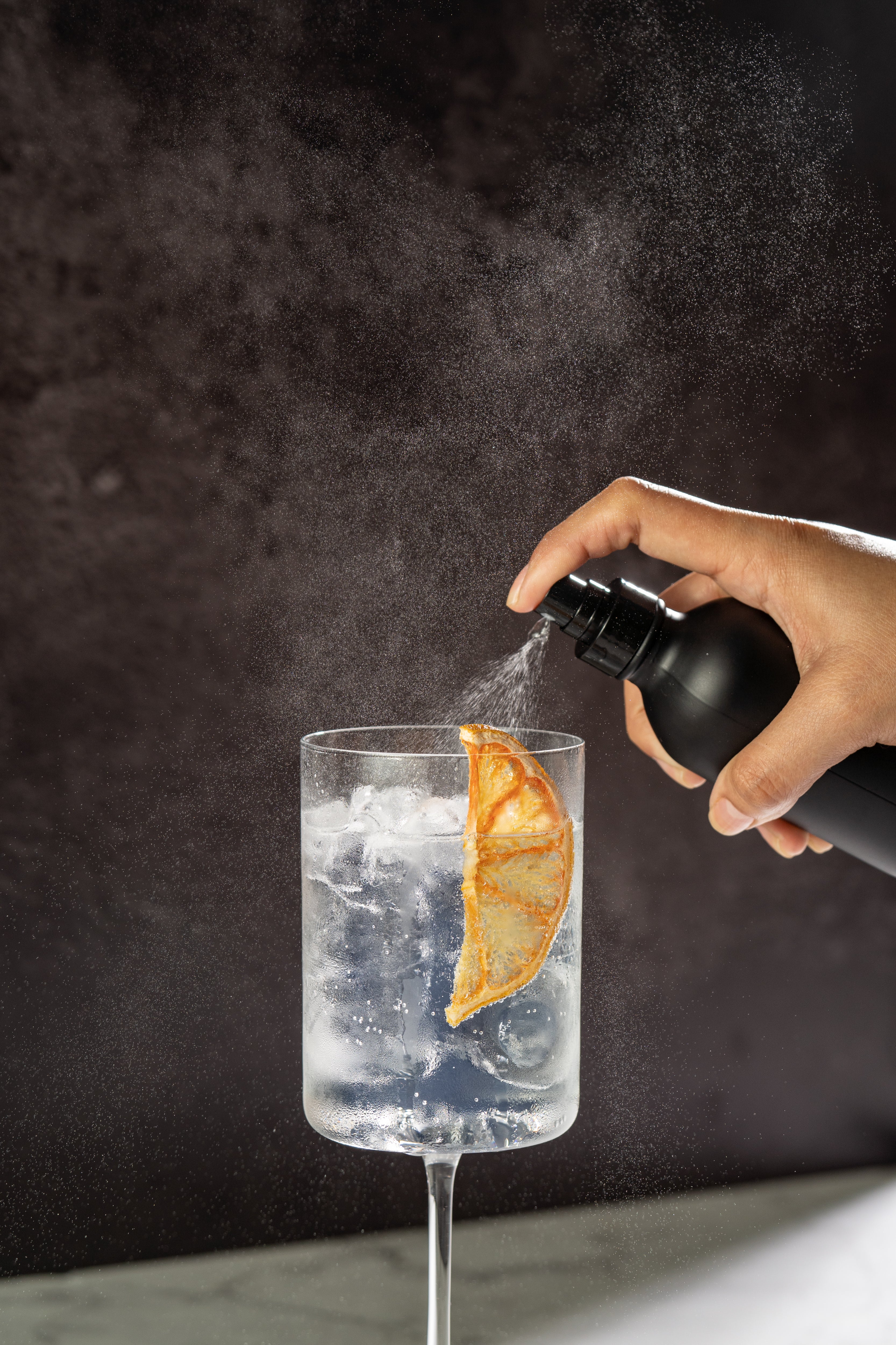 Hand spraying clear liquid from a black atomizer bottle into a tall square wine glass with clear liquid, ice, and dried orange garnish