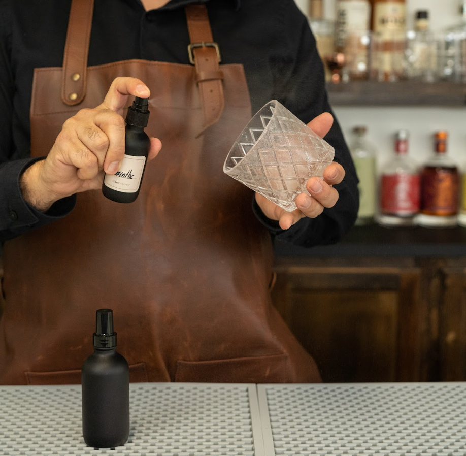 Bartender hand spraying clear liquid from a black atomizer bottle into a rocks glass at a pro bar, with another black bottle nearby