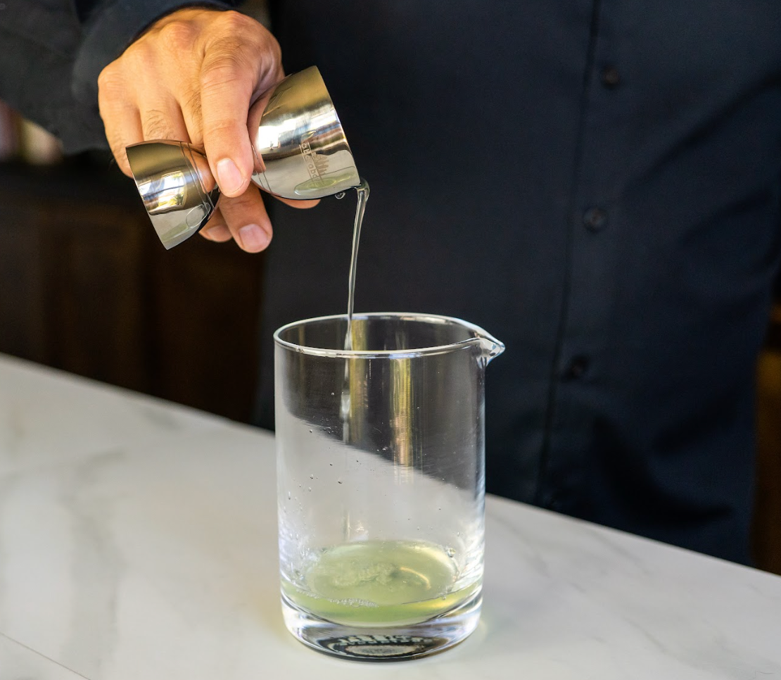 Hand pouring liquid from a stainless steel bell jigger into a clear cocktail mixing glass, on a white marble bartop