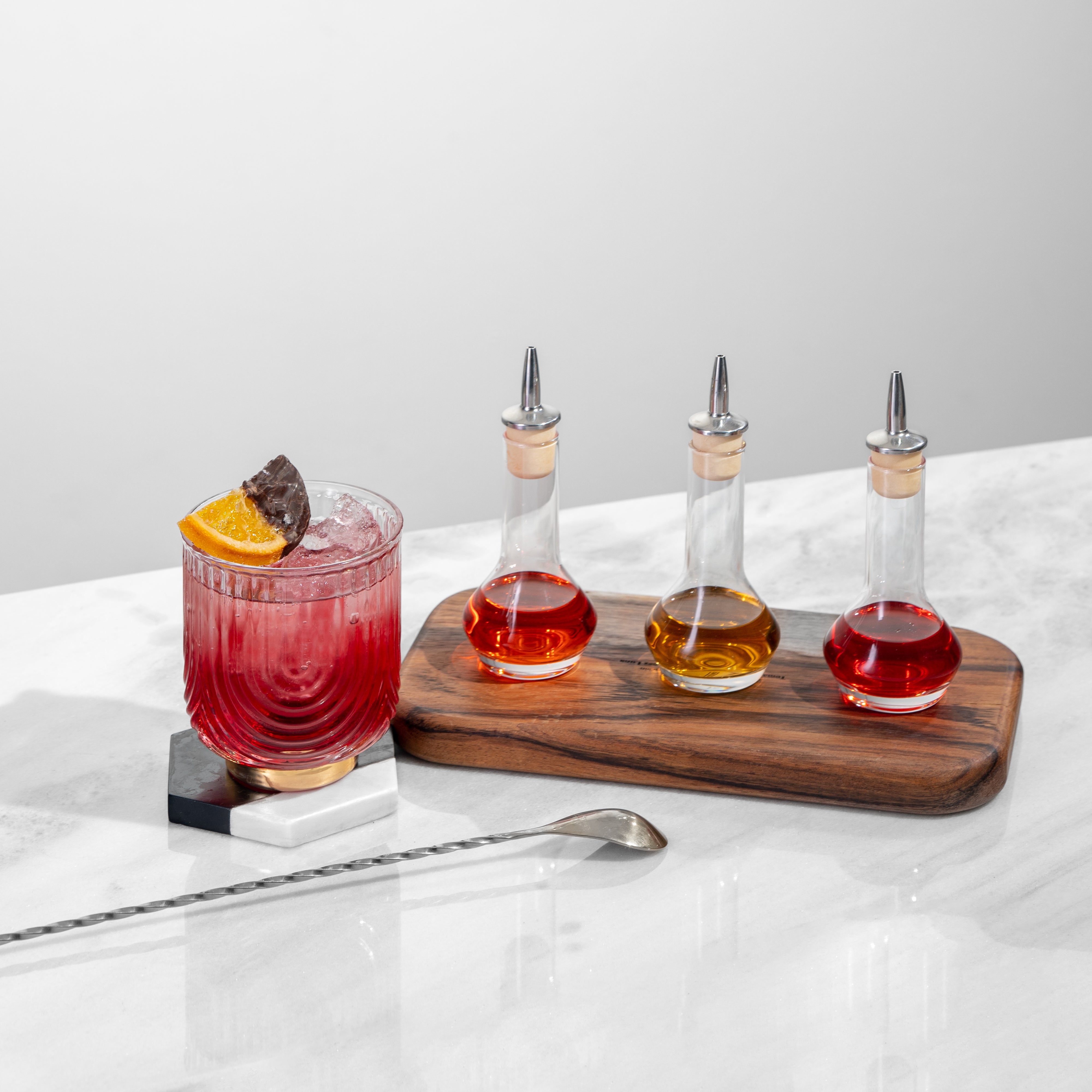 3 clear bitters bottles on a wood tray, each with red or amber liquid, next to a red cocktail with orange garnish & silver bar spoon