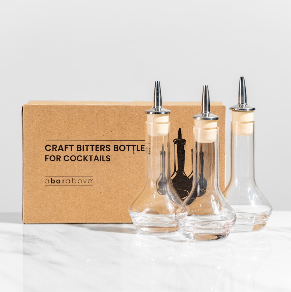 3 empty bitters bottles with round bases, sitting in front of their gift box which reads "craft bitters bottles for cocktails"