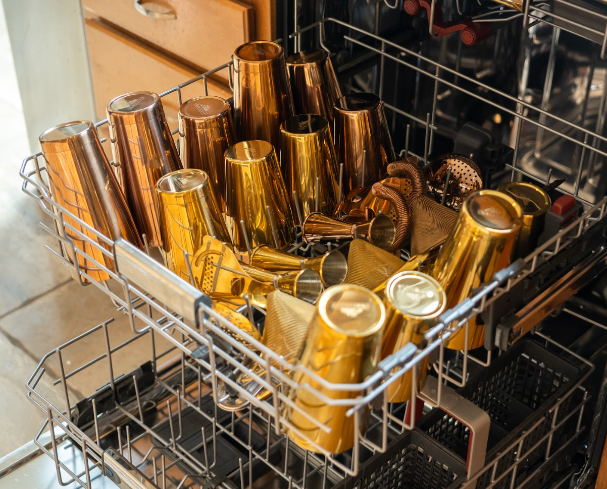 A dishwasher top rack full of gold and bronze cocktail shakers, jiggers, and strainers