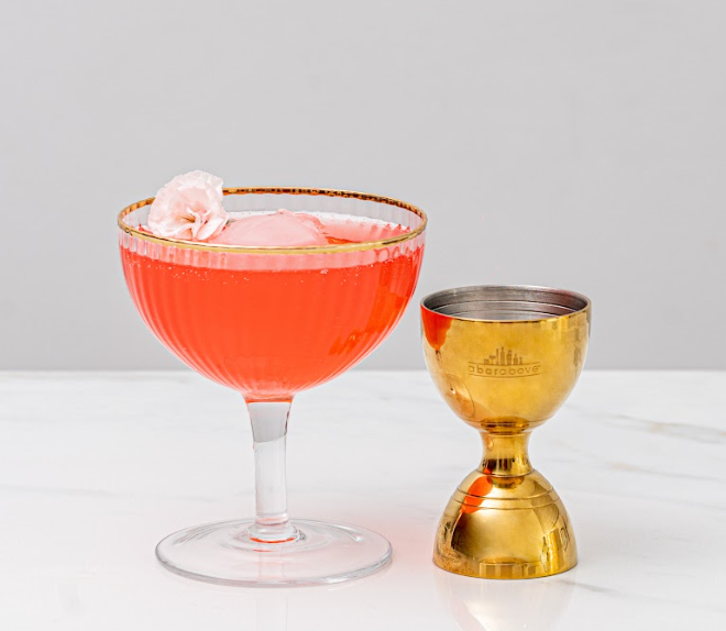 Pink-orange cocktail in a coupe glass with a pink flower garnish next to a gold bell cocktail jigger, standing on white marble