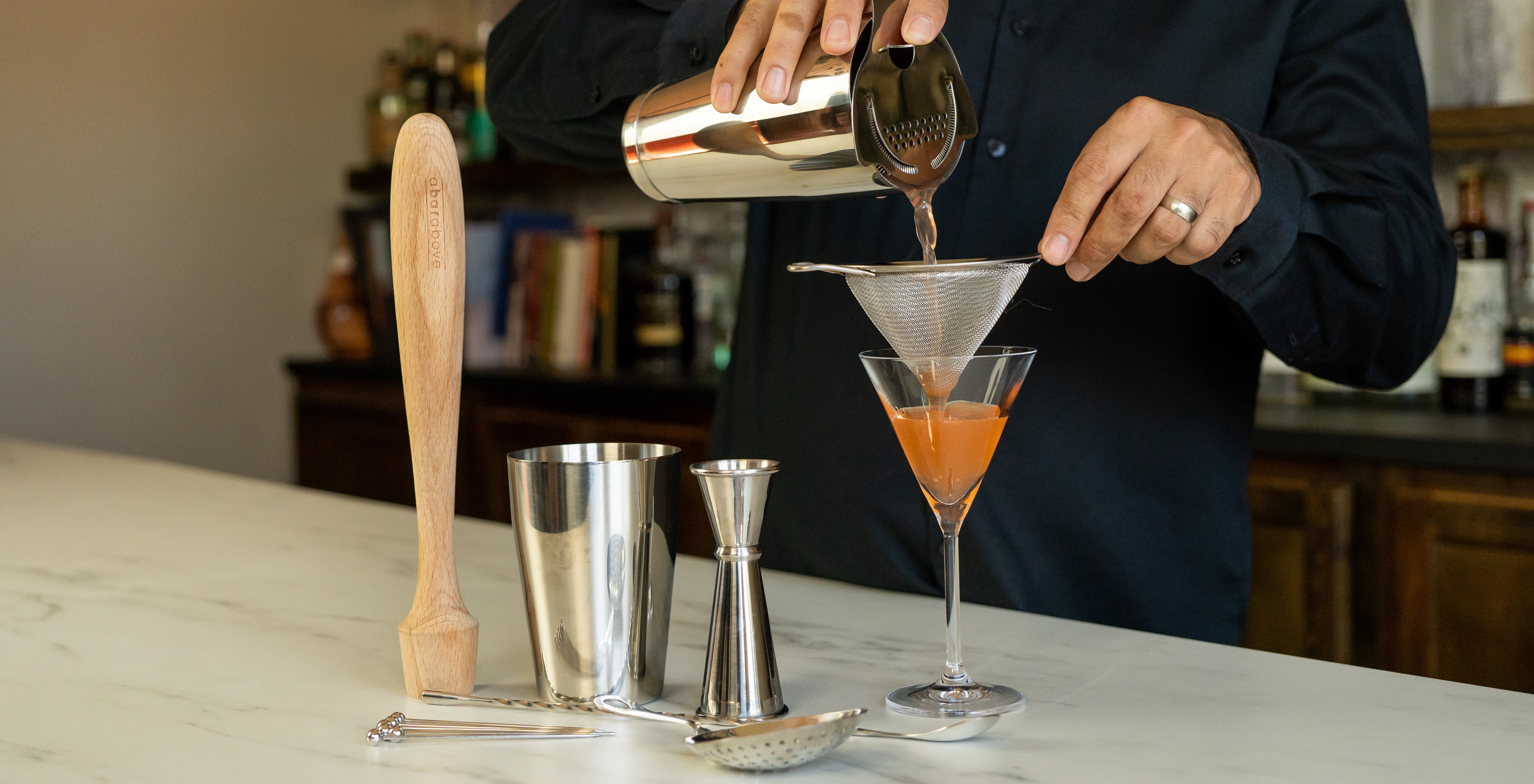 Bartender's hand pouring an orange cocktail from a cocktail shaker through a fine strainer into a Martini glass, with bar tools next to it