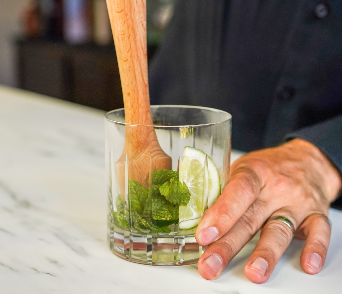 Close-up of a hand holding a rocks glass with mint and lime being muddled with a wood cocktail muddler