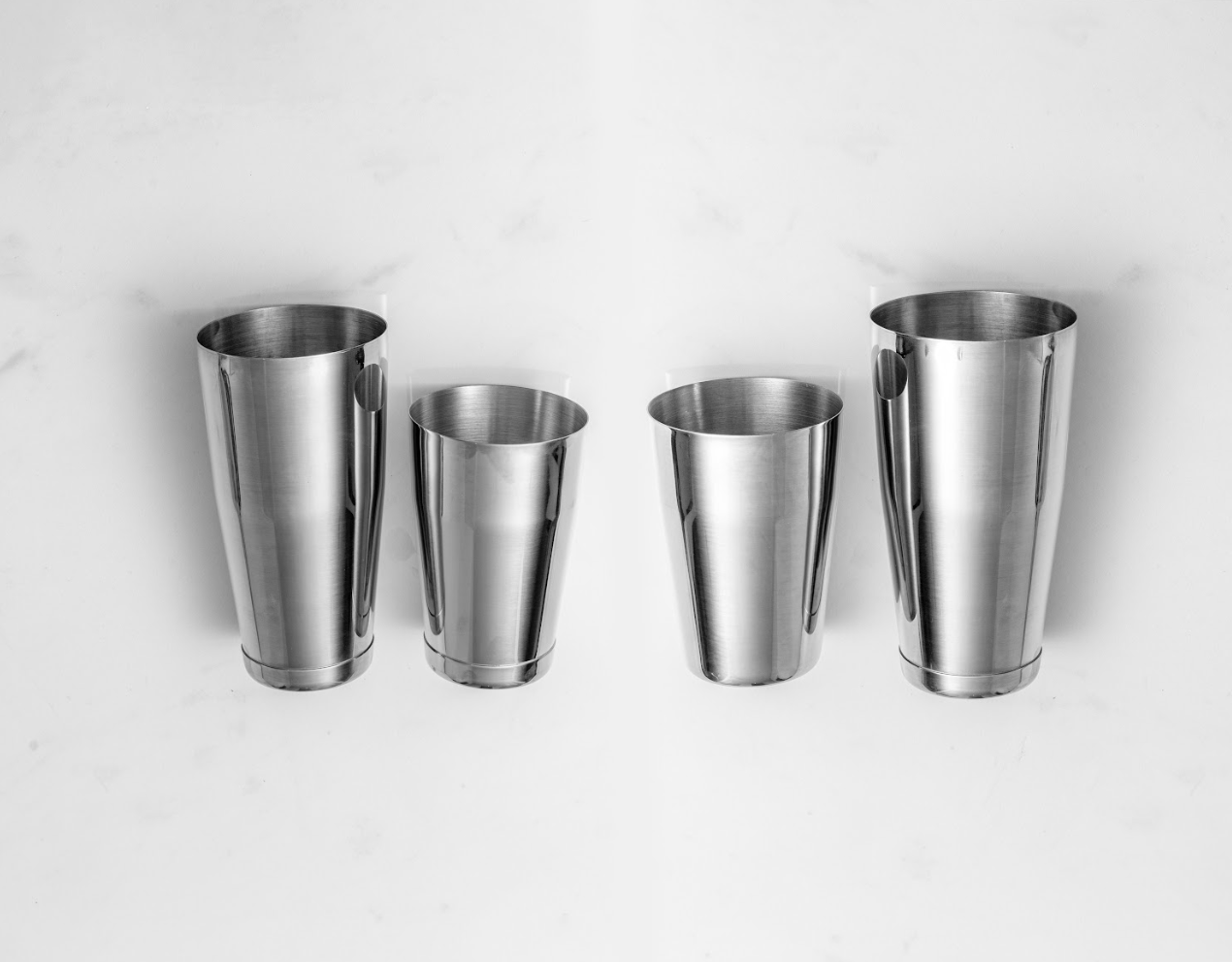 Two sets of Boston cocktail shakers lying flat on white marble. One set has circle weights on the bottom & one set has only 1 weighted cup