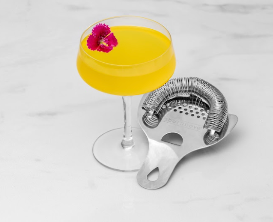 Yellow cocktail with fuchsia colored flower in a coupe glass, next to a stainless steel Hawthorne cocktail strainer on a marble counter