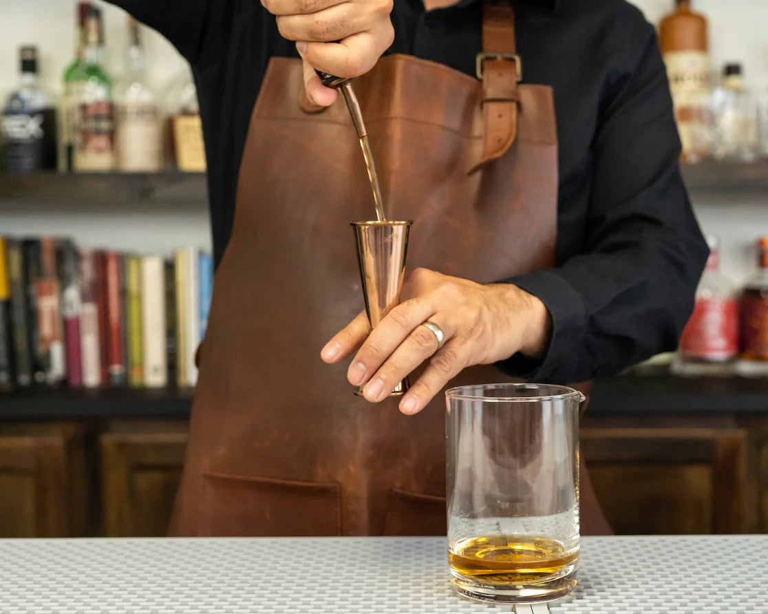 Hand pouring liquid from an bronze pour spout into a cocktail jigger behind a clear mixing glass with brown alcohol, in a pro bar
