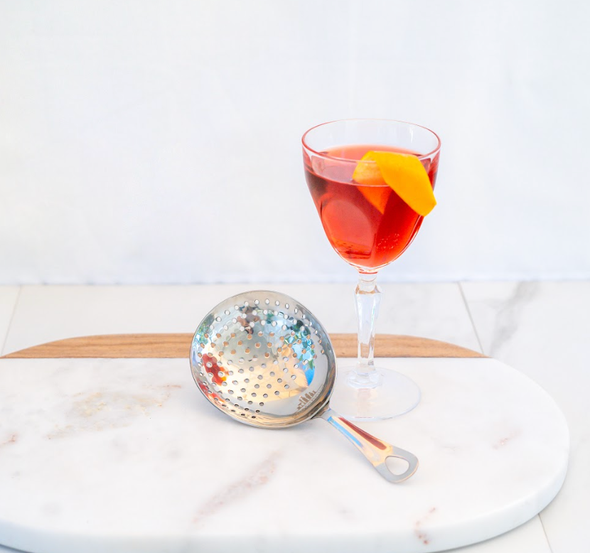 Red rocktail wtih orange peel garnish in Nick & Nora glass next to a stainless steel julep strainer, set on a white marble tray