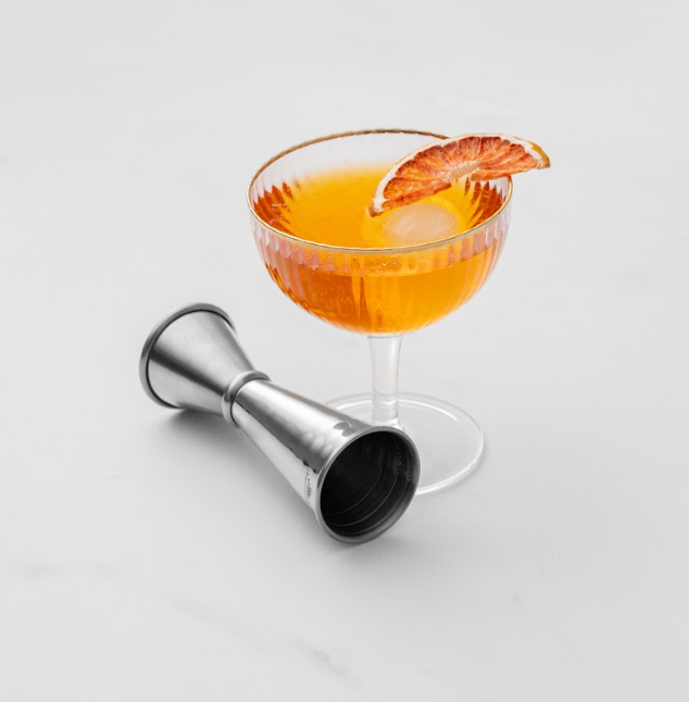 Orange cocktail in a coupe glass with dried orange garnish next to a stainless steel cocktail jigger on a white backdrop