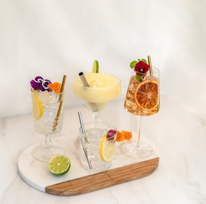 Margarita, orange cocktail in wine glass, and 2 clear cocktails in wine & rocks glasses, all with straws and flowers, on a white marble tray