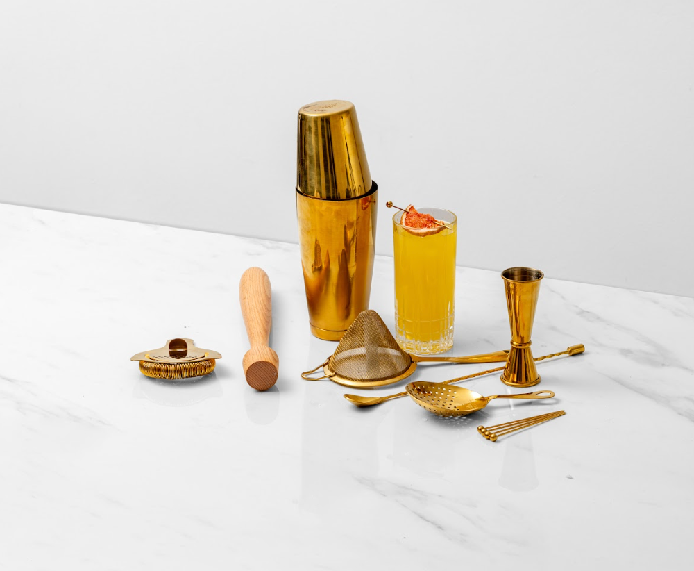 Gold cocktail shaker, 3 strainers, jigger, spoon, picks, & wood muddler, all around an orange cocktail in a tall glass with orange garnish