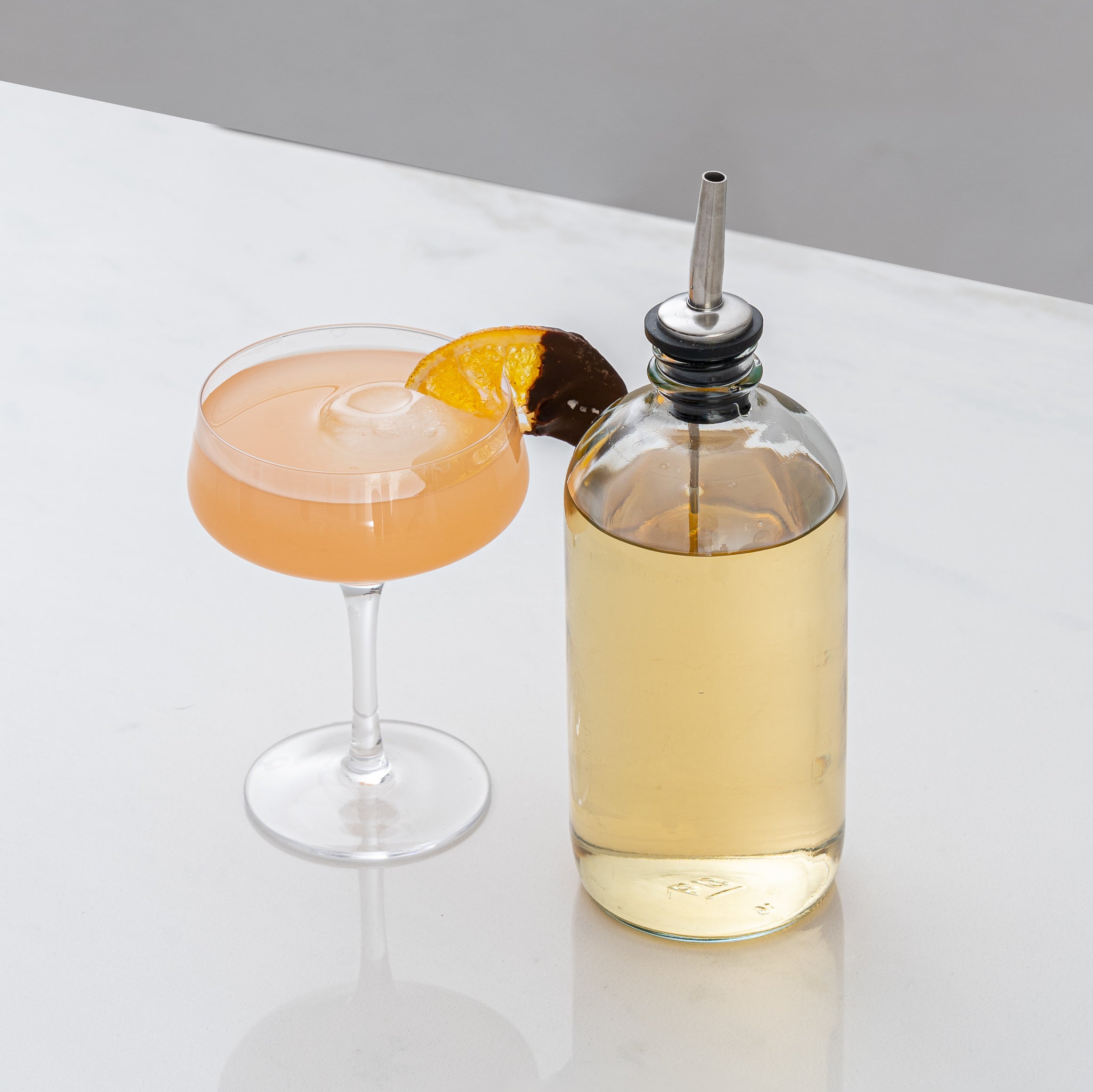 Peach colored cocktail in a coupe glass with orange slice, next to a bottle filled with golden liquid & an alcohol pour spout on it 