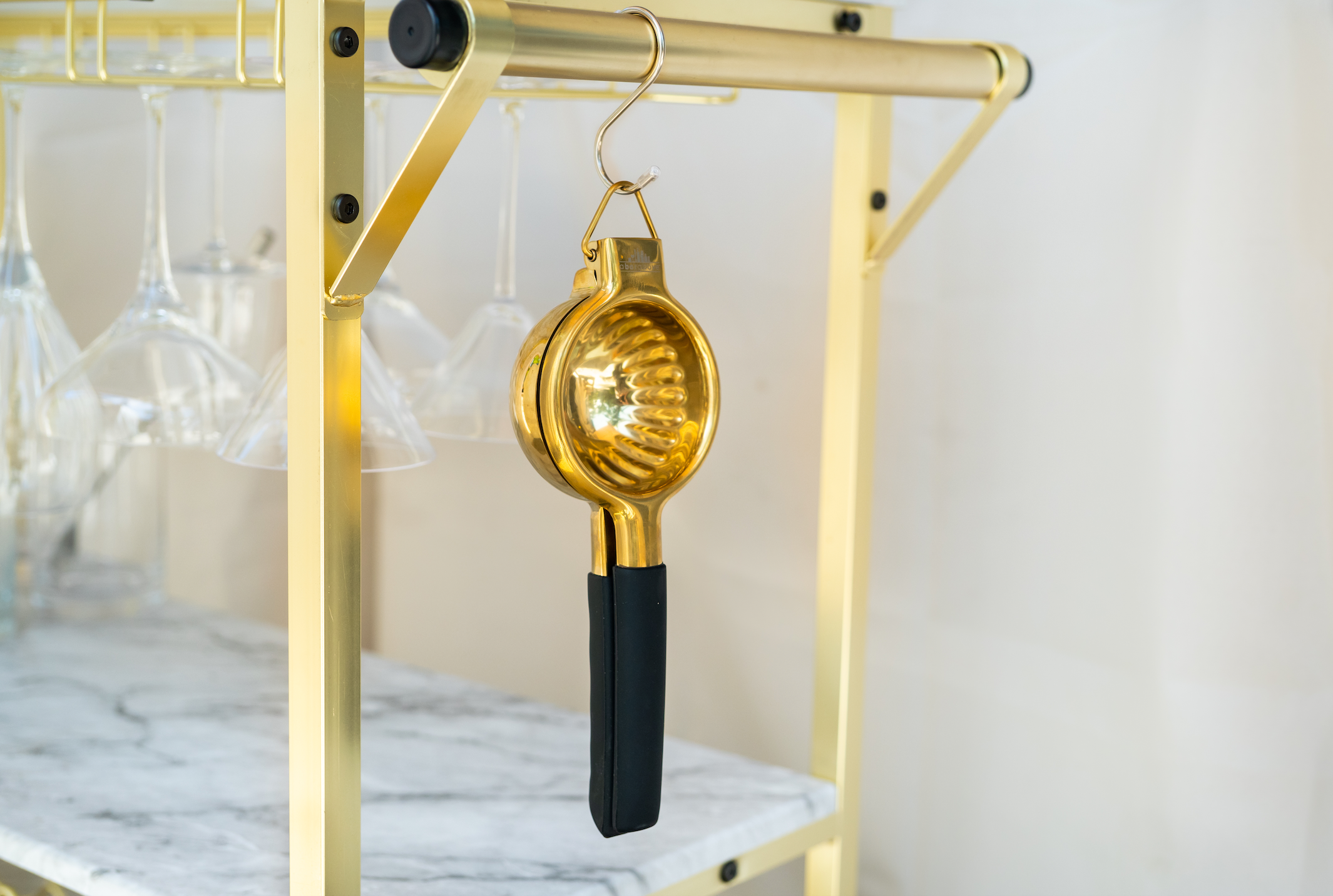 Gold lemon juicer hanging from a hook on a marble & gold bar cart with Martini and wine glasses hanging on it
