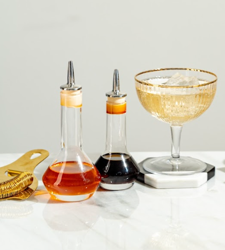 2 clear bitters bottles, 1 with red liquid and 1 with brown, next to a gold Hawthorne strainer and champagne cocktail in a coupe glass  