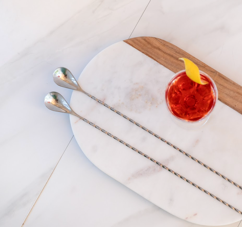 Top-down photo of a red cocktail with lemon peel garnish, sitting on a white marble tray next to 2 extra-long bar spoons with spiral handles