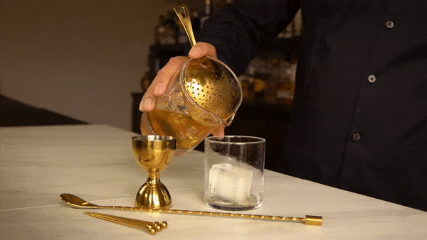 Hand holding a mixing glass with gold julep strainer, about to pour a drink into a rocks glass, next to jigger, spoon, and cocktail picks