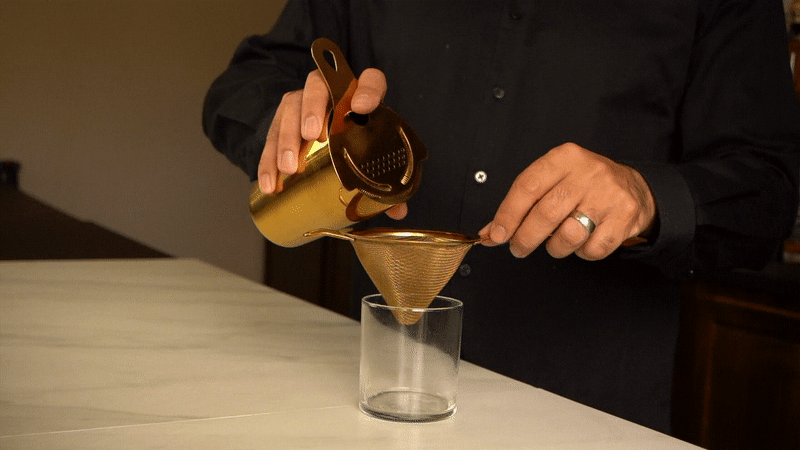Hand holding a gold cocktail shaker with Hawthorne strainer, about to pour a drink into a rocks glass through a fine strainer