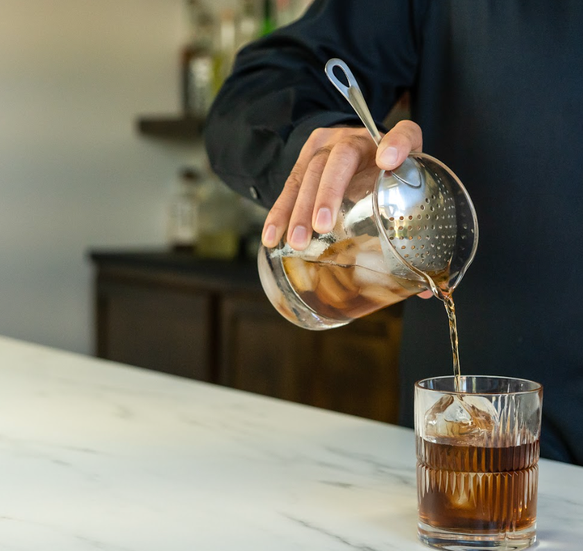 Hand pouring a brown cocktail into a rocks glass tumbler from a cocktail mixing glass with a julep strainer fit inside the top