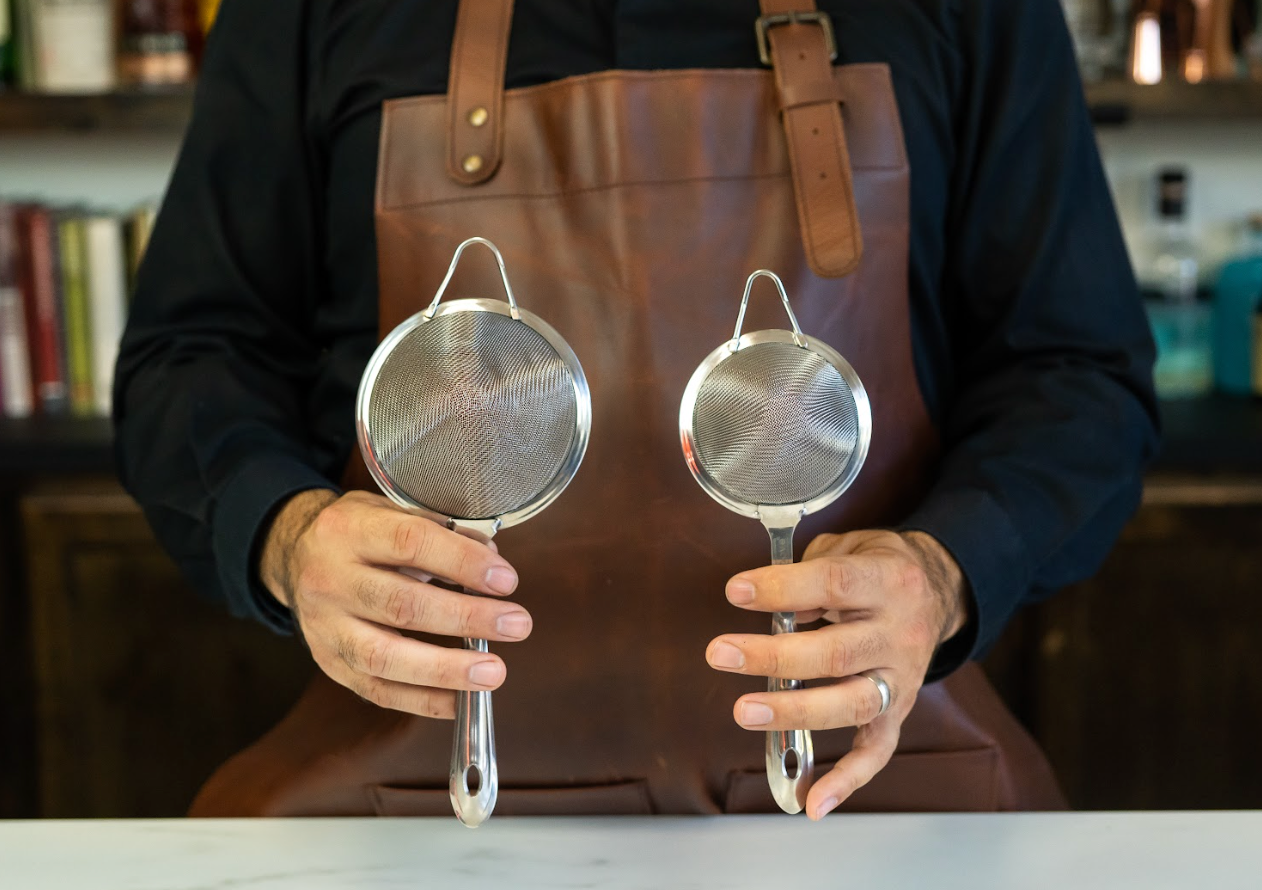 Bartender's hands holding up two sizes of stainless steel fine mesh strainers in a pro bar-- one larger and one smaller fine strainer 