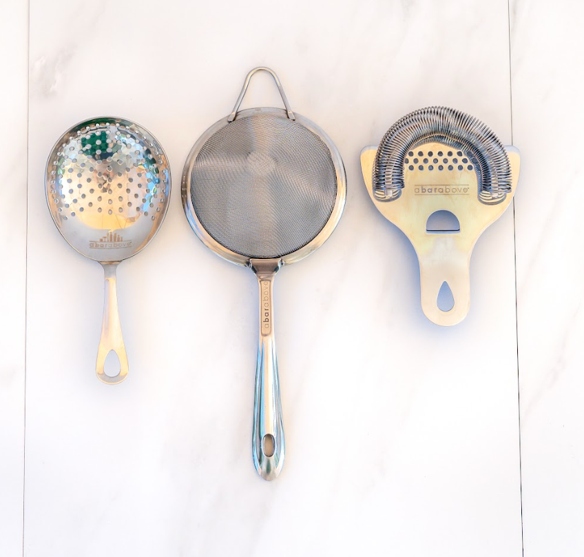 Stainless steel julep, fine mesh, and Hawthorne strainers lying flat on a white marble counter