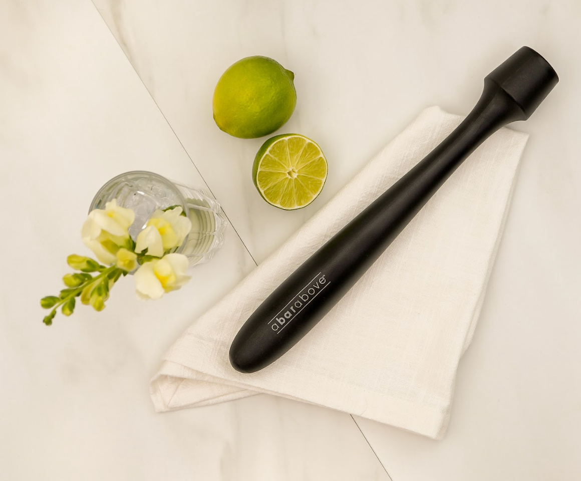 Flatlay photo of a black cocktail muddler on a white napkin, next to a vase of flowers, a whole lime, and a cut half of a lime