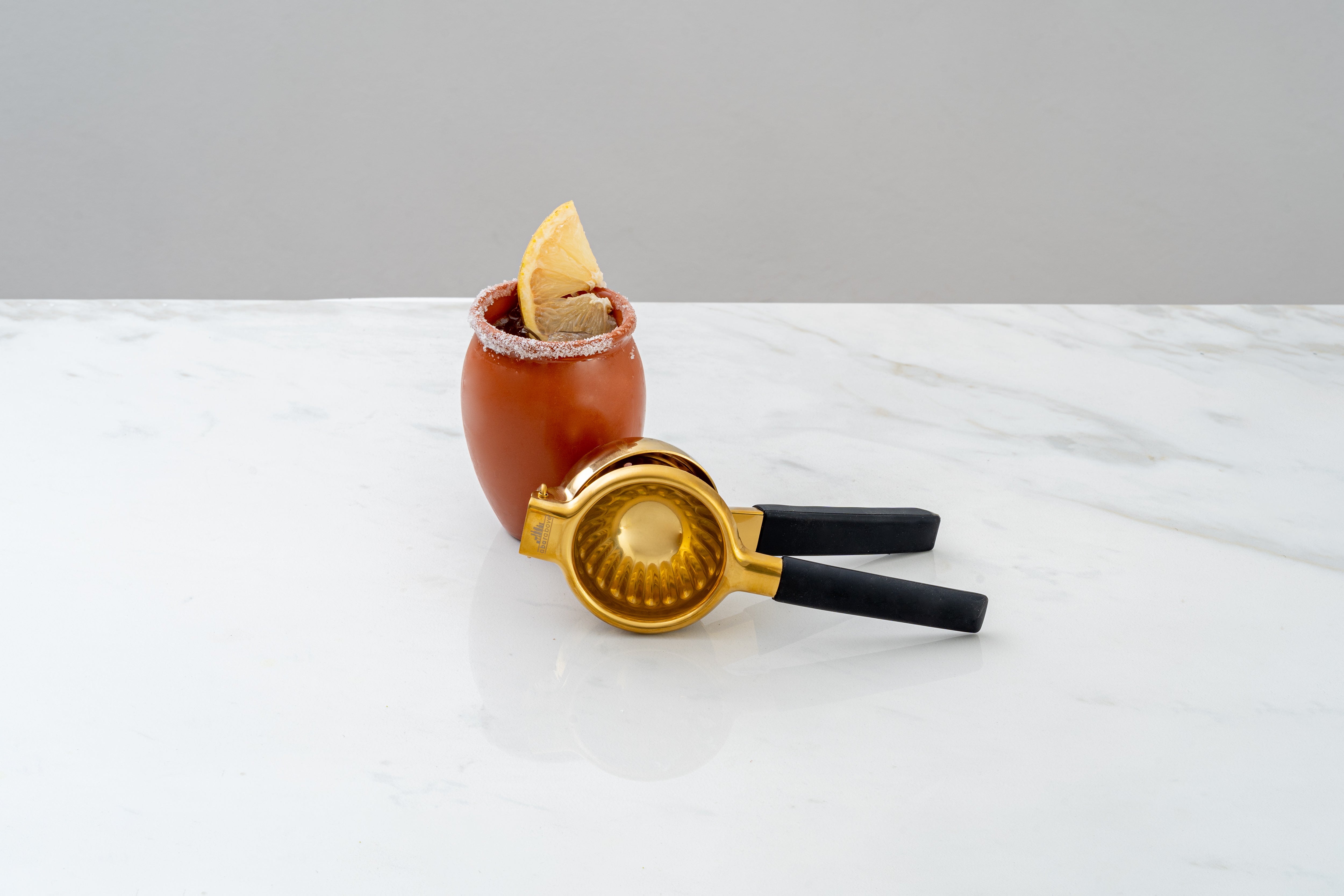 Brown vase-like drink cup with salt rim & lemon garnish, behind a gold citrus juicer with black handles, sitting on a white marble counter